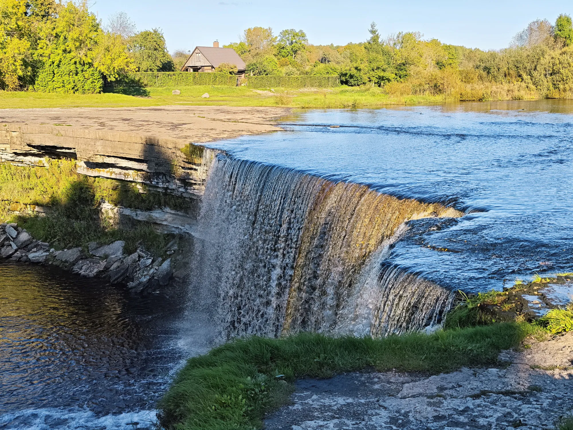 The Jägala waterfall in late afternoon light, a wide, low curtain of water pouring over a limestone ledge.