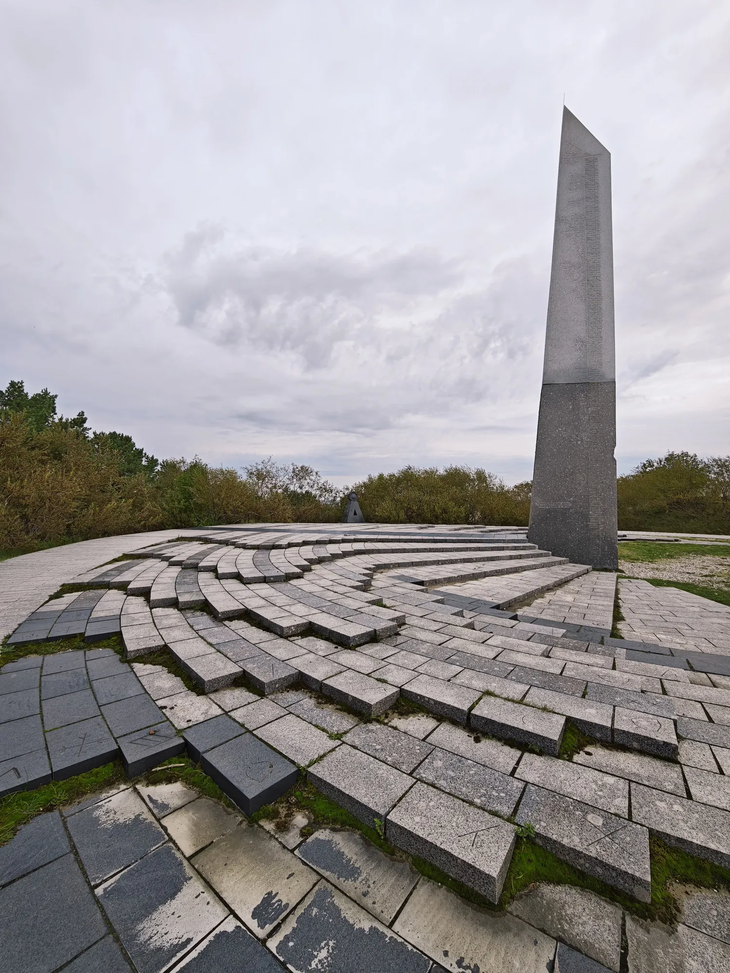 The granite sundial column on top of the Parnidis dune near Nida, under a heavy sky, with pine forest behind.